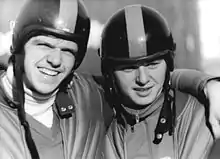 Two men wearing helmets embrace each other. The man on the left has his left arm over his partner's shoulder and grins while protecting his eyes from the direct sunlight with his brows.