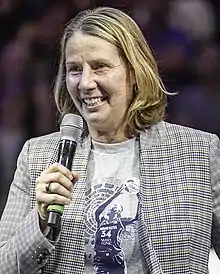 Portrait of a woman in her mid fifties holding a microphone with shoulder length highlighted hair, wearing silver hoop earrings, a Sylvia Fowles tribute t-shirt, gray-patterned suit jacket
