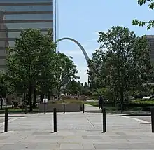 Four black bollards sit in the foreground, trees are visible in the middle, and in the distant background, a tall grey arch peeks out from behind office buildings.