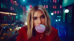 Dua Lipa blowing a bubble while sitting on a car in a traffic jammed city block.