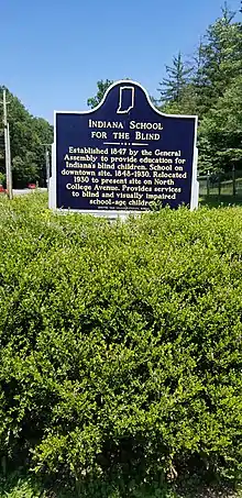 A historical marker surrounded by a bush with small purple flowers.  The sign reads, "Indiana School for the Blind. Established 1847 by the General Assembly to provide education for Indiana's blind children.  School on downtown site, 1848-1930.  Relocated 1930 to present site on North College Avenue.  Provides services to blind and visually impaired school-age children.