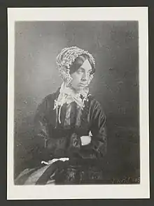 Valentine Delessert in a dark dress and white bonnet sitting with arms crossed staring away