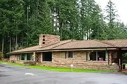 Brown one-story modern flagstone building, parking lot in front and fir trees behind it.
