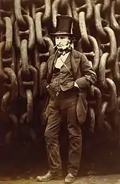 A 19th-century man wearing jacket trousers and waistcoat, hands in pockets, cigar in mouth, wearing a tall stovepipe top hat, standing in front of giant iron chains on a drum.
