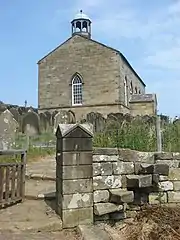 A small stone church standing in a churchyard on top of a hill.  On the near gable is a bell-cupola.