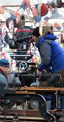 A man wrapped in a blue jacket and black hat looks down the scope of a large film camera. There is red, white, and blue bunting hanging overhead.