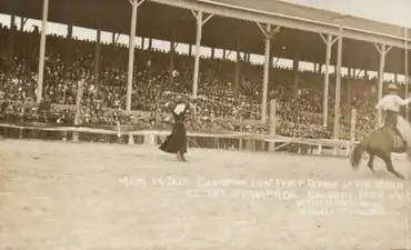 Woman in black dress roping a horse and rider in front of a grandstand.. Miss La Due Champion Lady Fancy Roper of the World at the Stampede. Calgary, Alta. 1912.