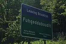 Highway sign with "Leaving Brooklyn" on a blue background, then on a green background a big "Fuhgeddaboudit", followed by "Mary Markowitz" "Borough President" and "Michael R. Bloomberg" "Mayor" in two columns with two lines each.